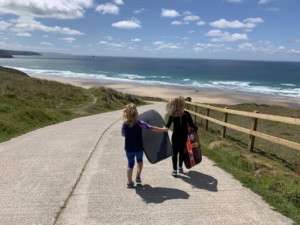 Children walking down hill to Perran Sands beach with body boards