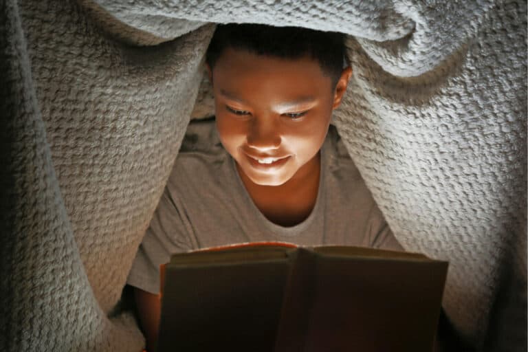 Boy reading a book by torch light under blanket