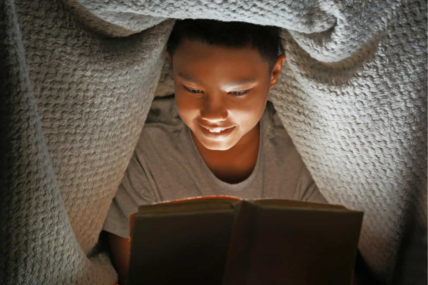 Boy reading a book by torch light under blanket