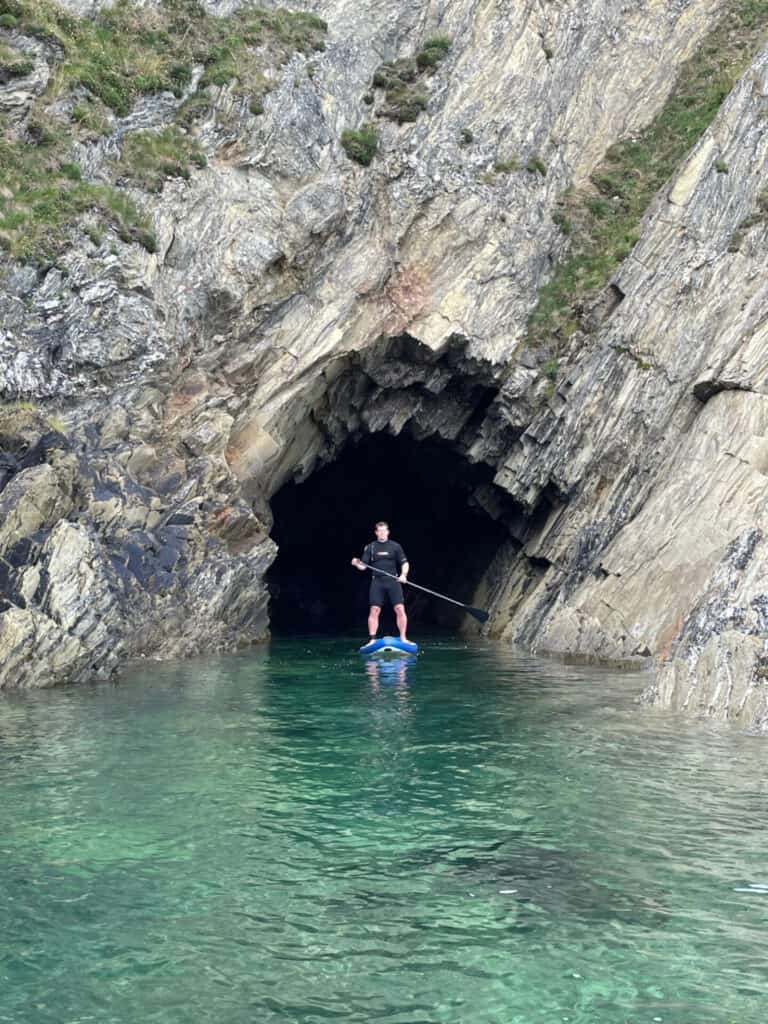 Mr Tin Box paddle boarding in front of cave at Maenporth Beach in Cornwall