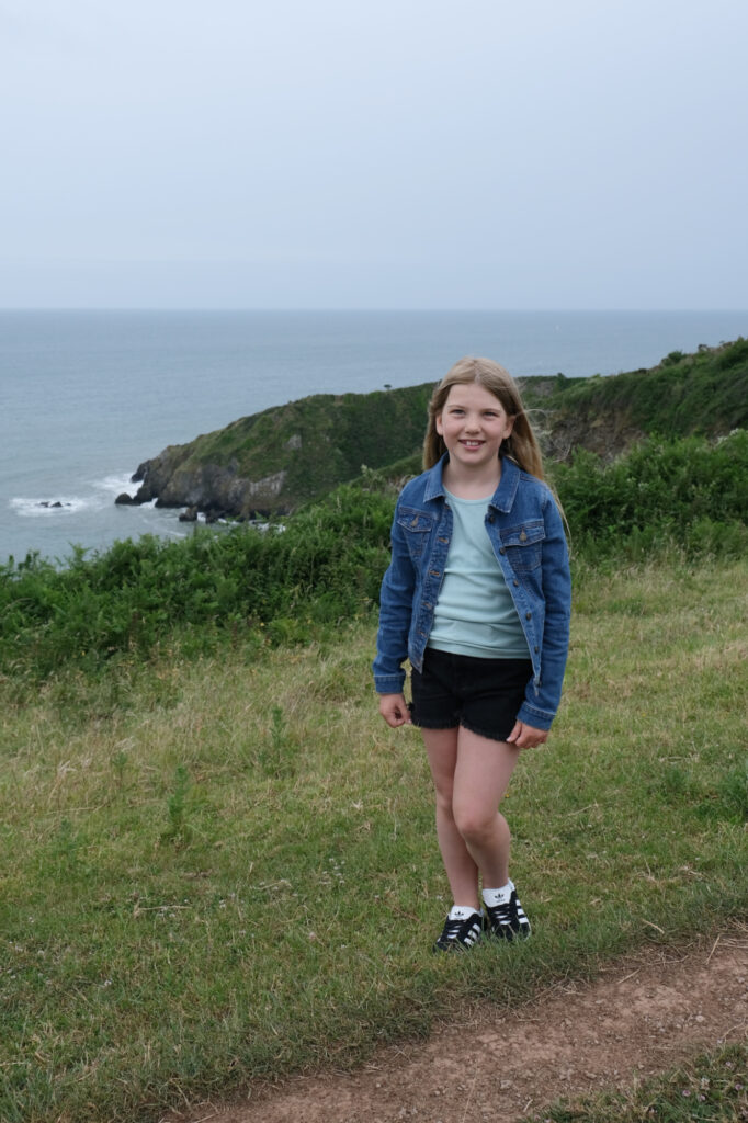 Girl in shorts, vest top and denim jacket in front of coast view