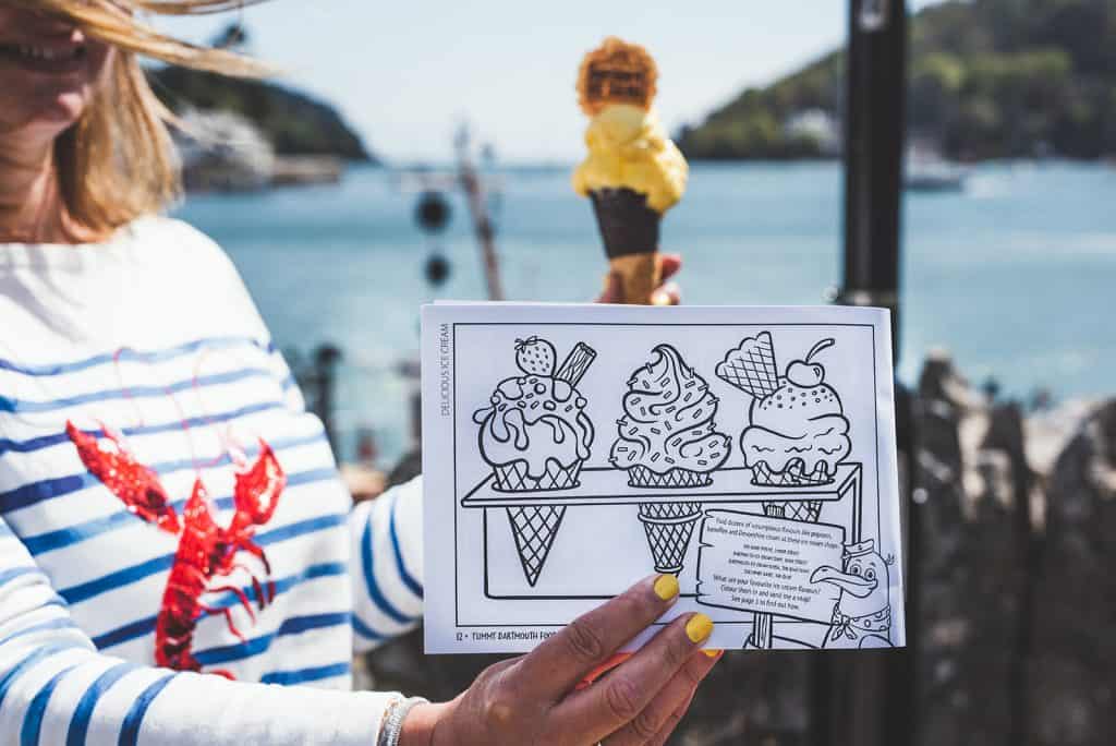 Claire holding an ice cream and colouring book open at a page with ice creams to colour in. In the background is a view of the Rive Dart in Dartmouth, Devon