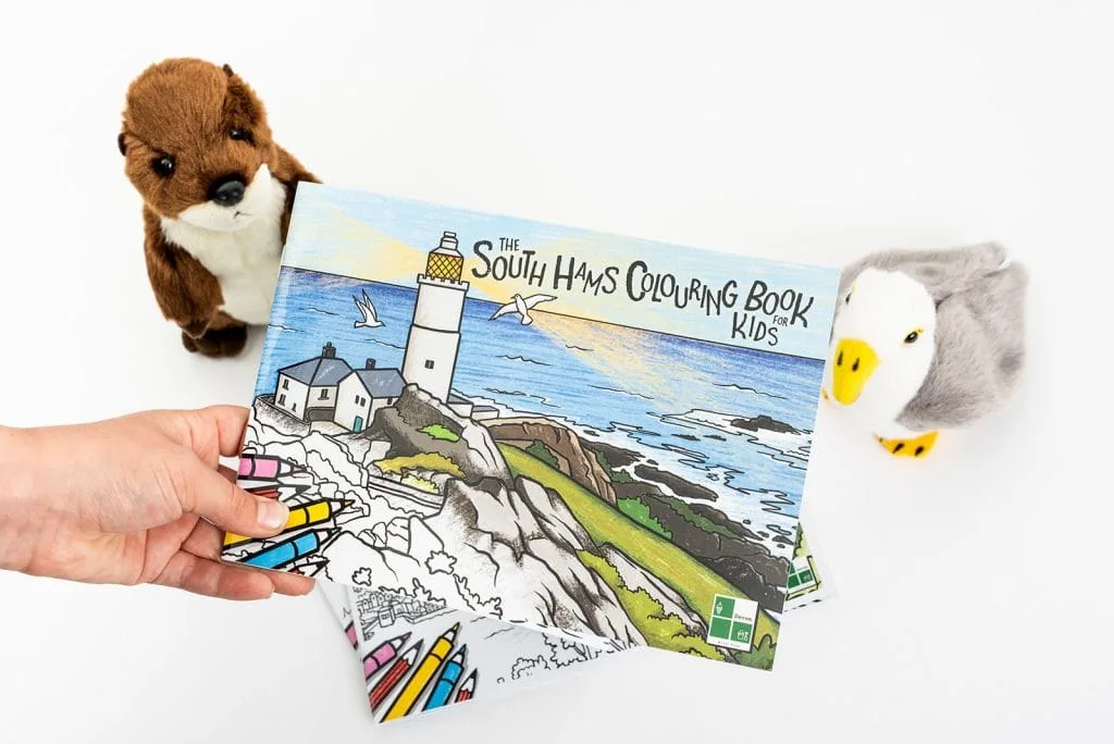 A hand holds up a copy of the South Hams colouring book. on the cover is a coloured in drawing of Start Point Lighthouse in South Devon