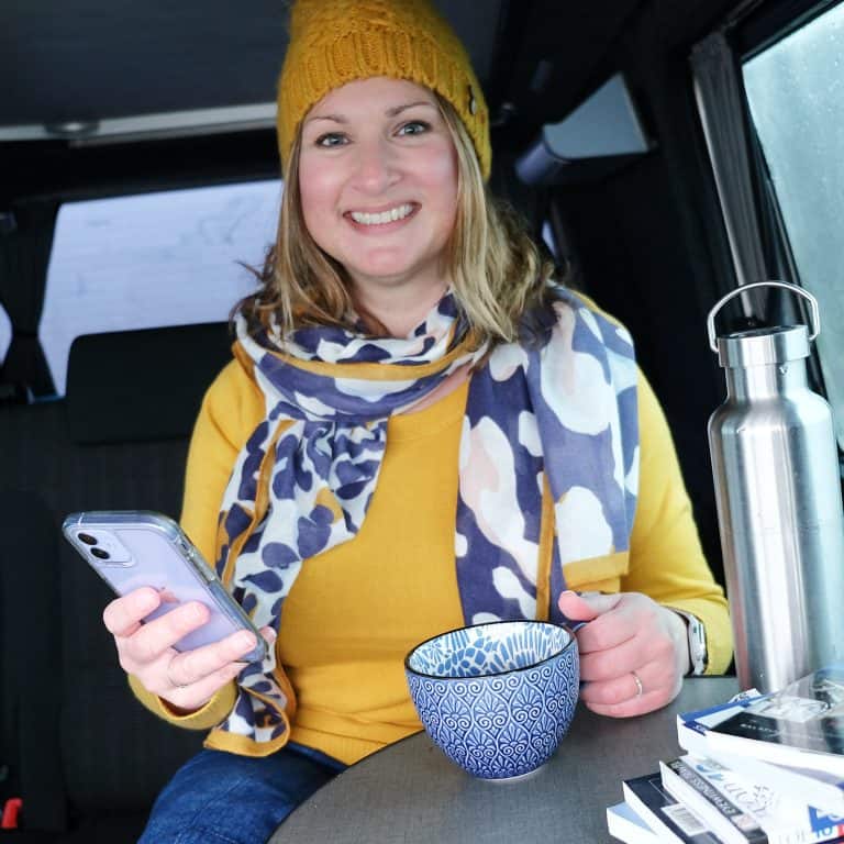 Claire Hall - award-winning travel blogger - holding phone and mug while sat in camper van