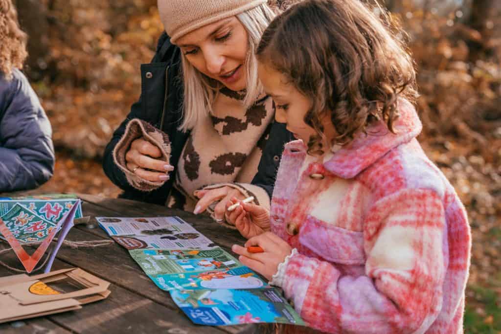 Woman and child in winter clothes complete activity book on picnic bench in forest