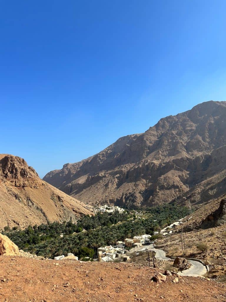 View down the Wadi Tiwi canyon towards a cluster of houses and plantations below. The wadi road winds up the cliffs