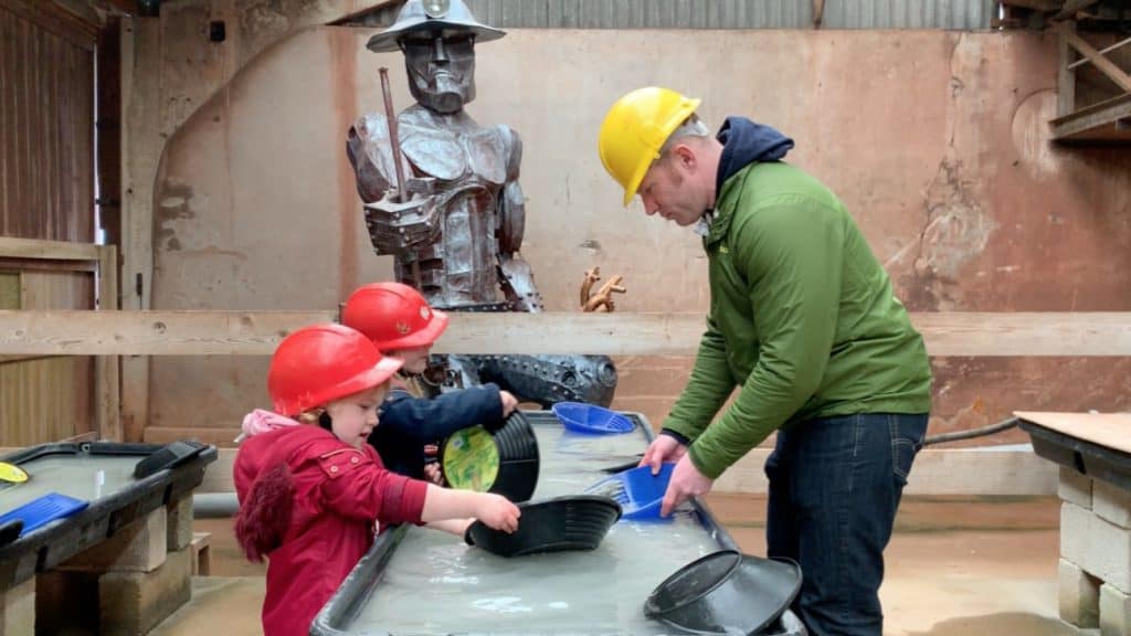 Family panning for gold in water trough at Geevor Tin Min with tin sculpture of miner in background