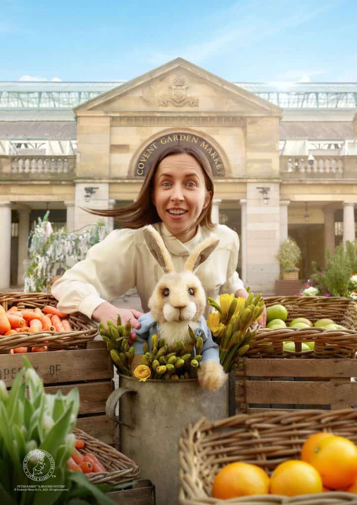 Woman smiles at camera with Peter Rabbit puppet in fruit and vegetable boxes. Covent Garden is in the background
