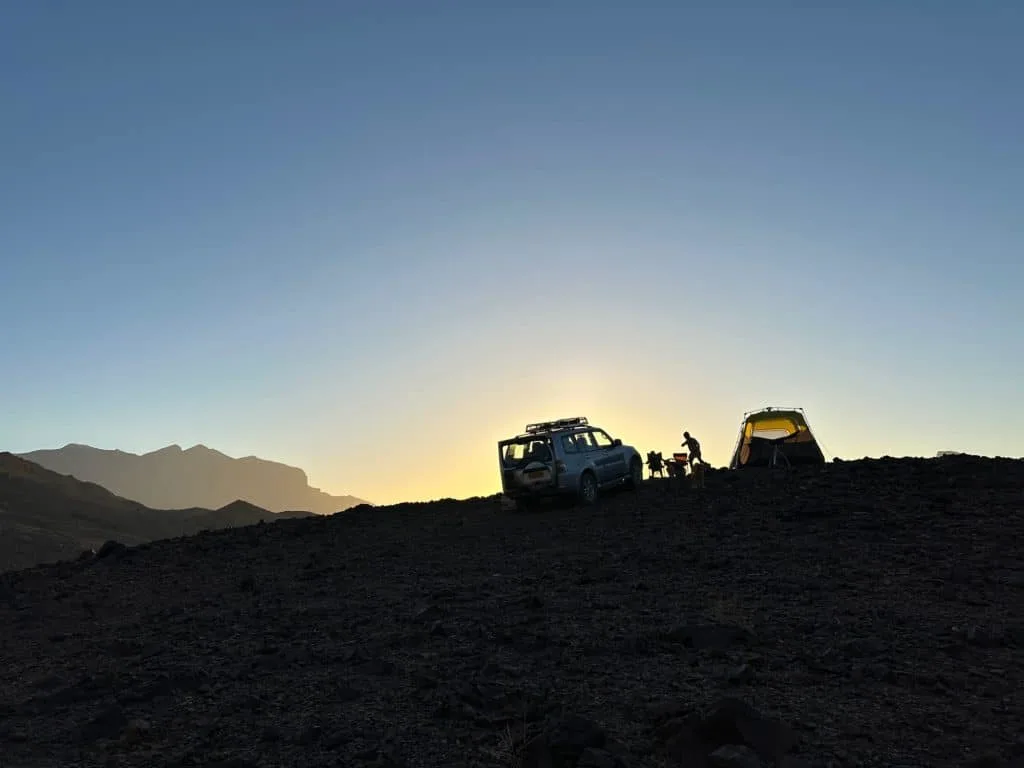 Outline of a 4x4, tent and people on the top of a rocky outcrop at dusk