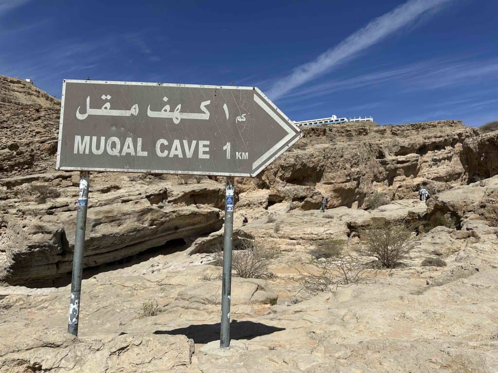 A brown sign on poles with 'Muqal Cave 1km' written on it in Arabic and English