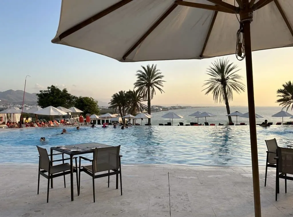 Sunset view of the outdoor pool at the Crown Plaza Muscat. In the foreground there is an umbrella, table and chairs. People are splashing in the pool. In the background you can see Shati al Qurum beach