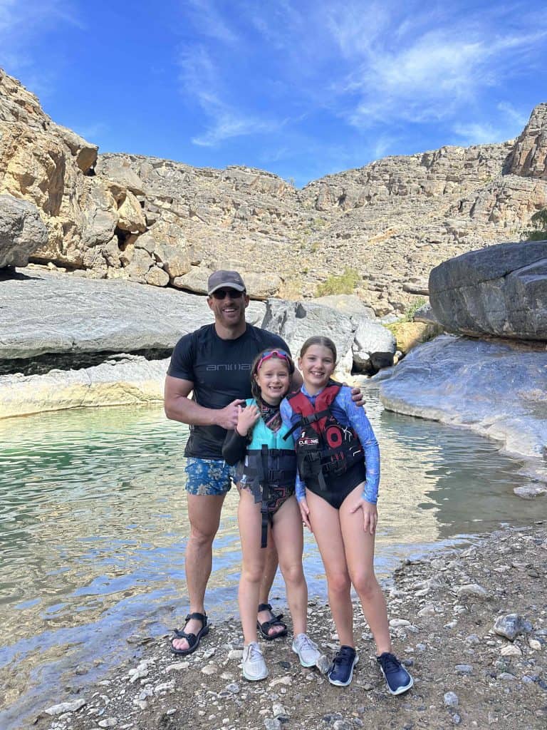 Mr Tin Box and daughters stand beside the water in Wadi Damm wearing conservative swim wear. The girls wear buoyancy aids