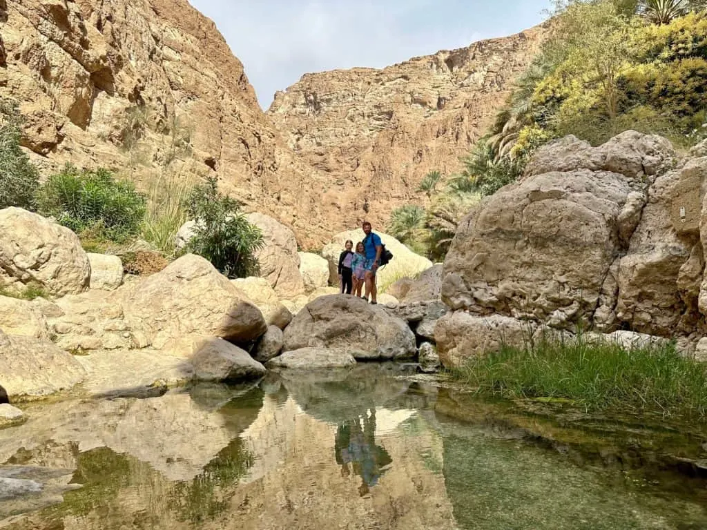 Father and daughters stood in front of pool of clear water in the rocky valley of Wadi Shab