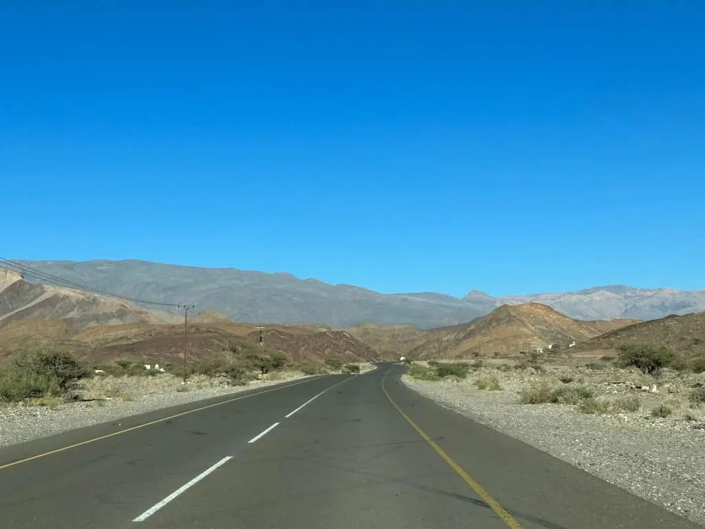 A tarmac road stretching out into the distance with mountains ahead and scrub land to either side. Some of the rocks include layered rock in zig zag formation