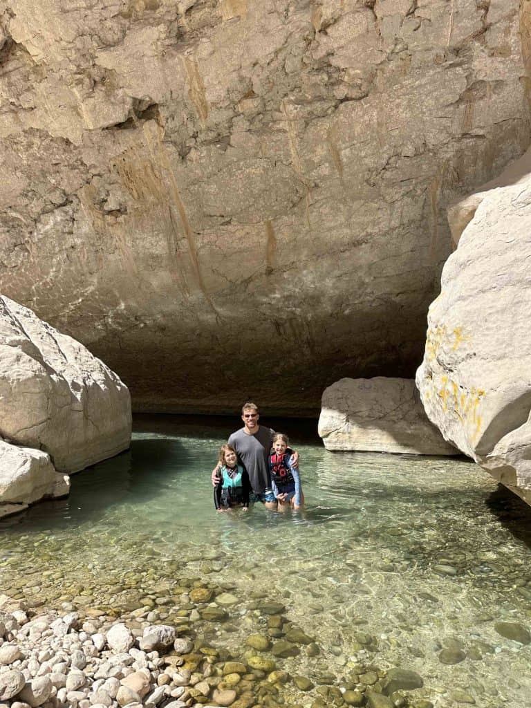 Mr Tin Box and daughters standing knee deep in the clear waters of Wadi Bani Khalid with large boulders towering above them