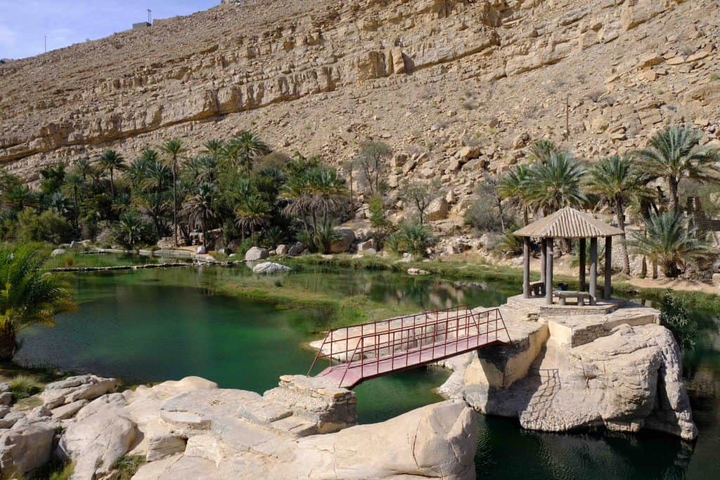 View of the first pool at Wadi Bani Khalid with a bridge and canopy in the foreground. The wadi water looks clear and inviting. The sand coloured valley wall rises in the background