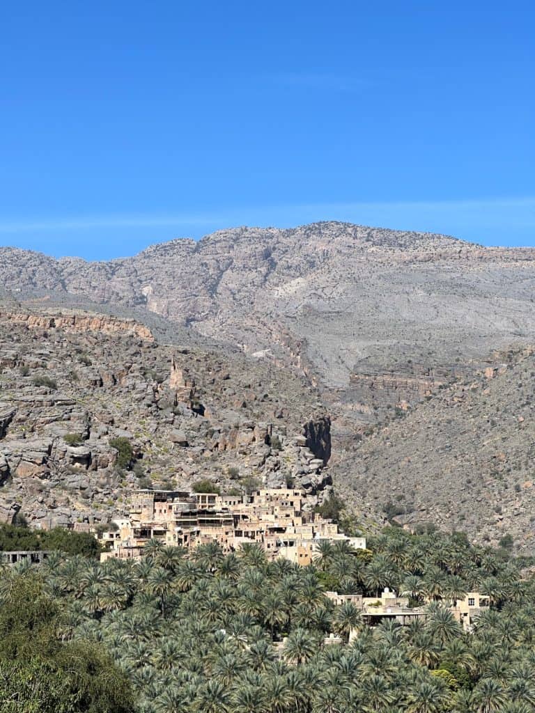View of the sand coloured houses of the historic village Misfat with palm groves below. The village sits on the side of a mountain