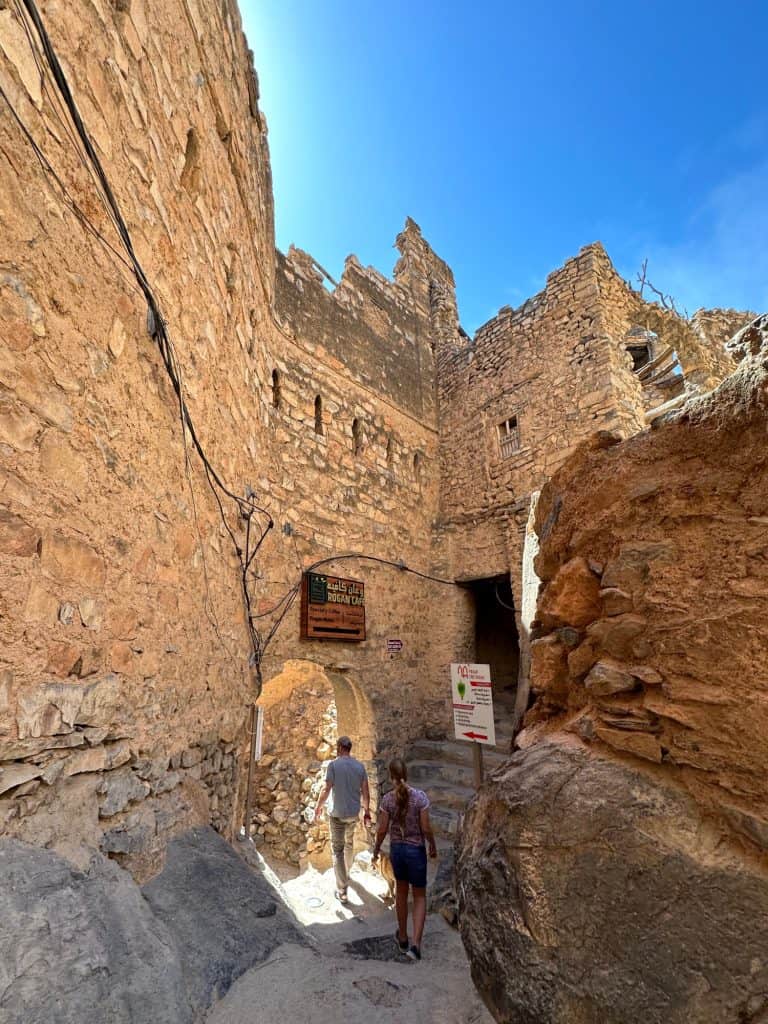 Allan and daughter walk through archway in ancient sandstone village walls at Misfat