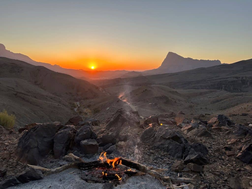 Sunsetting over the foothills of Jebel Shams in Oman's Hajar Mountains. A campfire is in the foreground and the white villas of a town can be seen in the distance