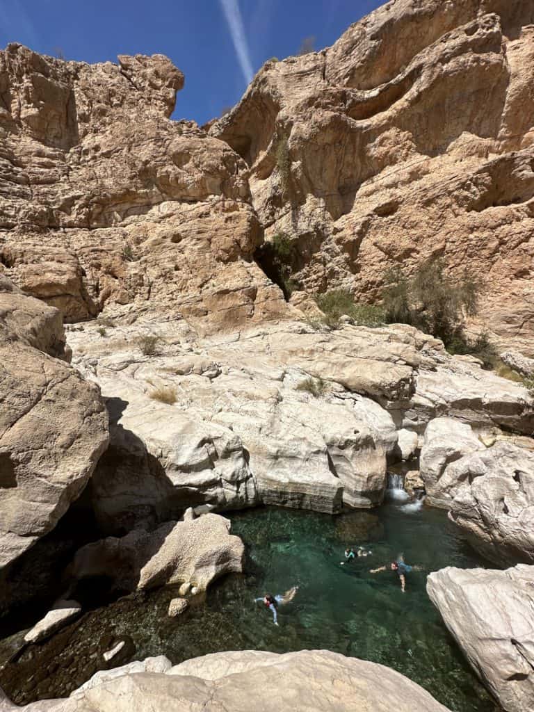 Three people can be seen swimming in the turquoise waters of Wadi Bani Khalid below. There are rocky cliffs all around