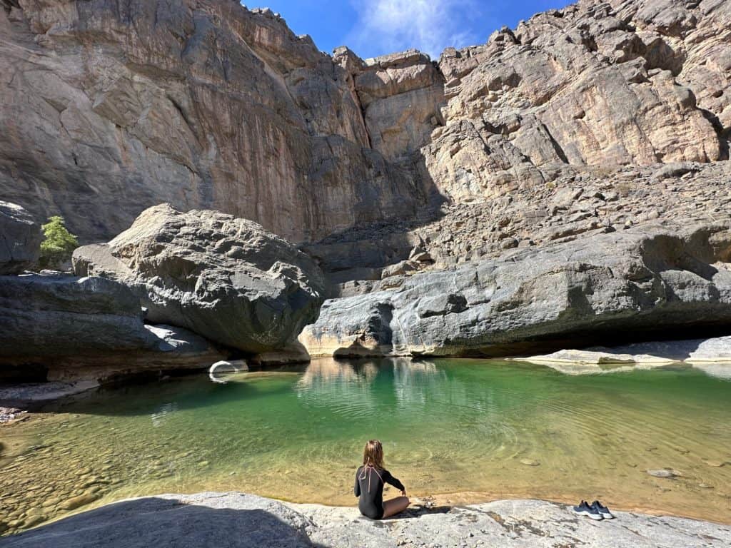 Child sat throwing stones into turquoise pool of Wadi Damm with valley wall rising up in front of her