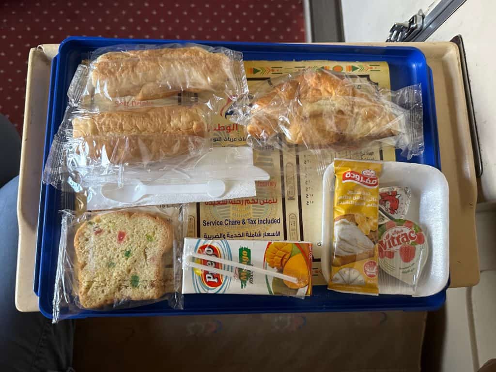 Breakfast on the sleeper train including bread rolls, a piece of cake, juice box and croissant with jam and cheese laid out on a tray