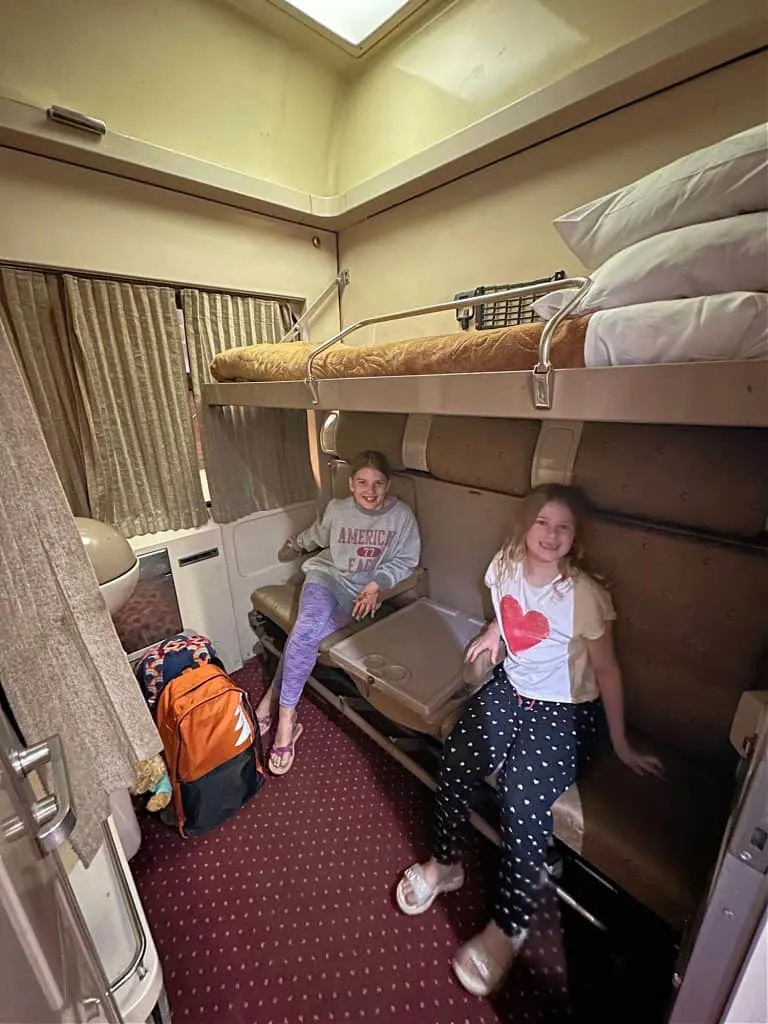 Two girls sat on on the seat in their sleeper train cabin. They are smiling. Above them the top bunk is made.