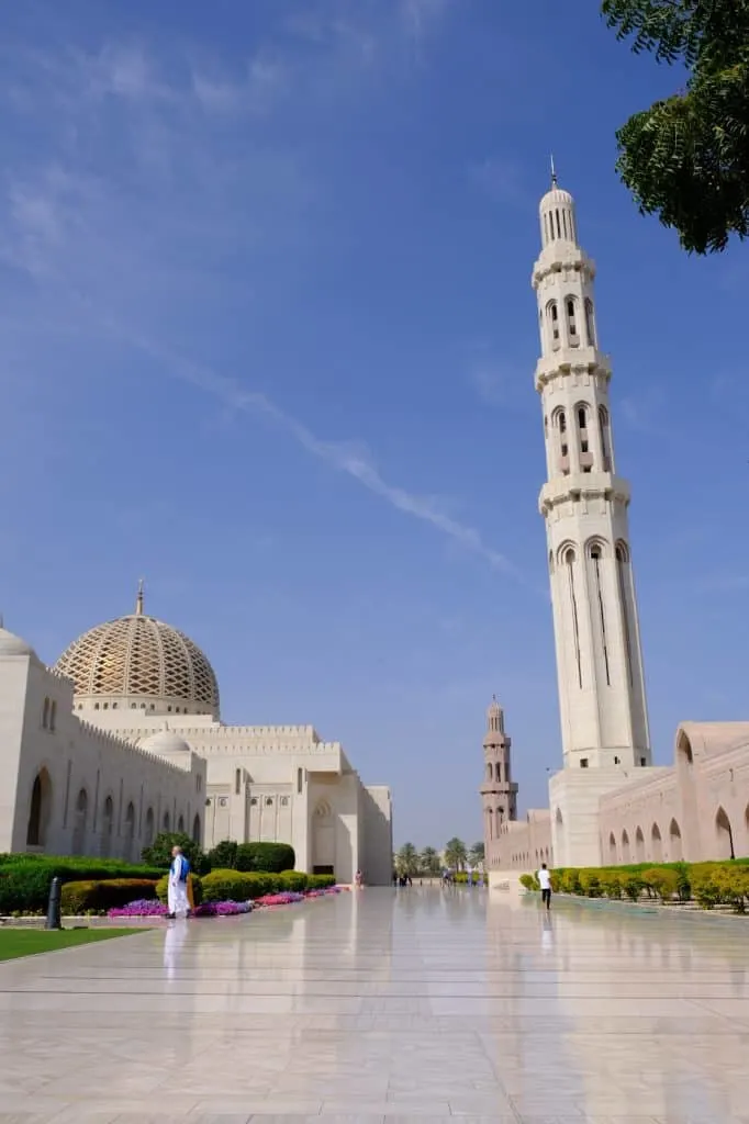 Exterior of the marble and sandstone mosque with rows of bushes in front. To the right is the tallest minaret and to the left is the dome of Sultan Qaboos Grand Mosque