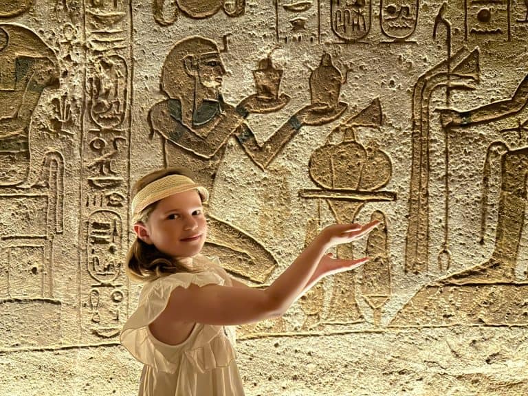 Child imitating an Egyptian in a hieroglyph behind her. She puts her hands our with palms facing up as if holding a jar