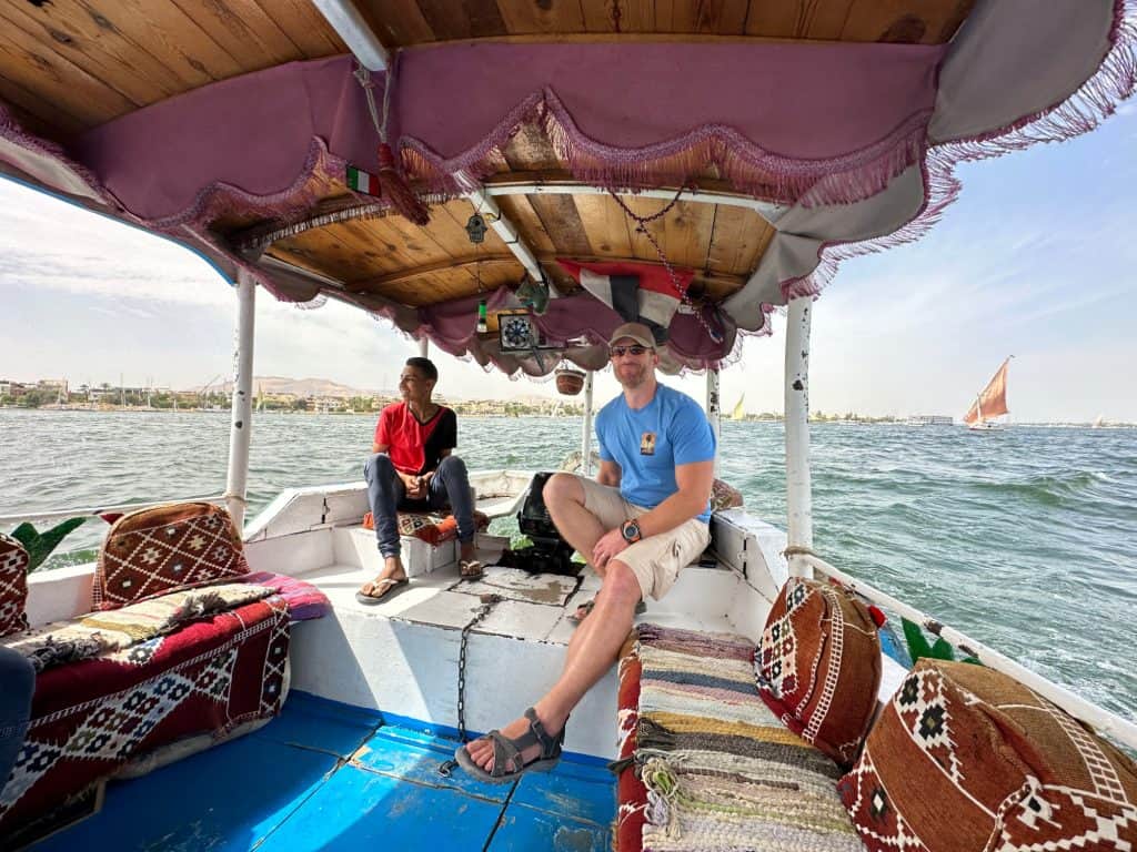 Mr Tin Box at the helm of our ferry across the nile. He sits beside the young driver. The boat has a canvas canopy and there is a view of the Nike behind