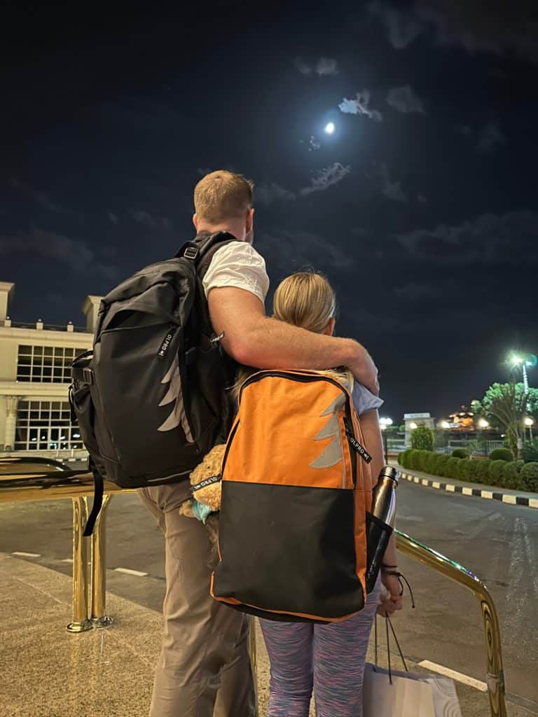 Mr Tin Box and daughter stand on steps of hotel looking at the moon in the night sky. They wear backpacks and the girl is holding a paper bag containing her breakfast