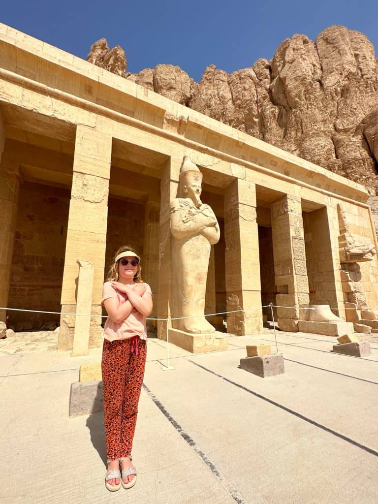 Girl posing with cross arms in front of a statue of Queen Hatshepsut at Deir Al-Bahari temple in Luxor