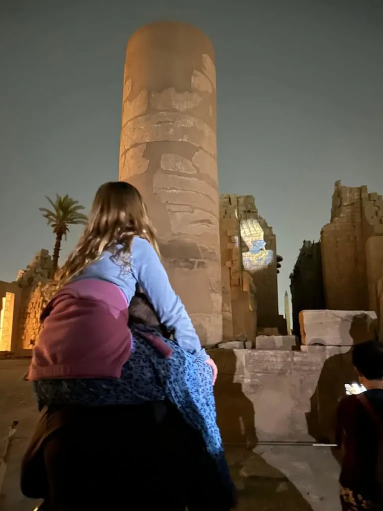 A projection of a pharaoh onto the wall of a temple at Karnak. In the foreground one of the girls is sat on Mr Tin Box's shoulders to get a better view. It is night time
