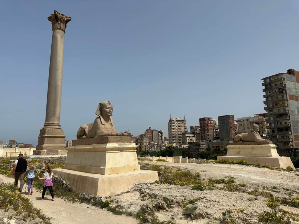 The family walk beside the a sphinx and the Pillar of Pompey at Serapeum in Alexandria. There are modern tower blocks to the right