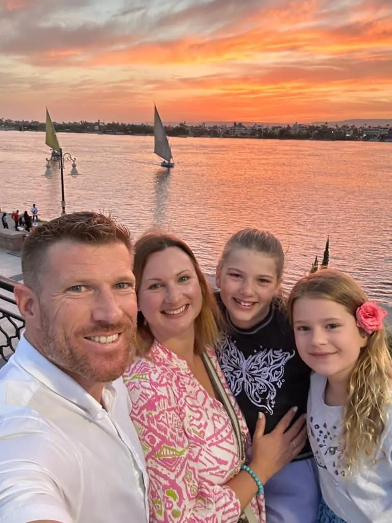 Tin Box family pose for a sunset selfie beside the Nile at Luxor. There are felucca sailing down the river behind them. The sky is pink and orange