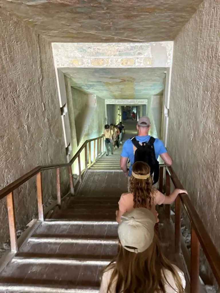 The family walk down the entrance tunnel of the tomb of Merenptah. There are hieroglyphs on the walls on either side. The entrance is steep and there are hand rails to help people descend