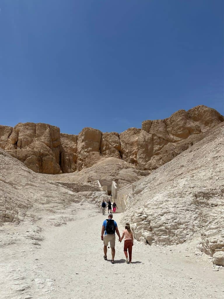 Mr Tin Box and daughter walk along a path through the Valley of the Kings. there are sandstone cliffs all around them and the sky is bright blue