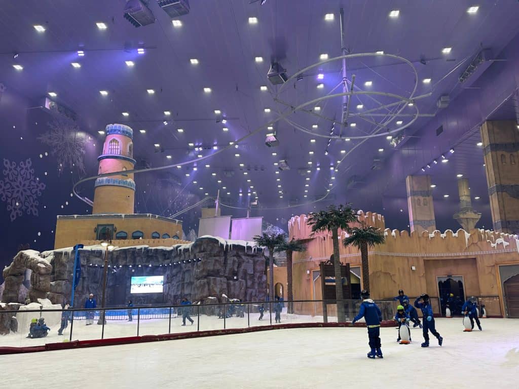 View of Snow Oman including Omani-style buildings covered in snow. An ice rink is in the foreground and an arial ride is suspended from the ceiling of the huge snow park room