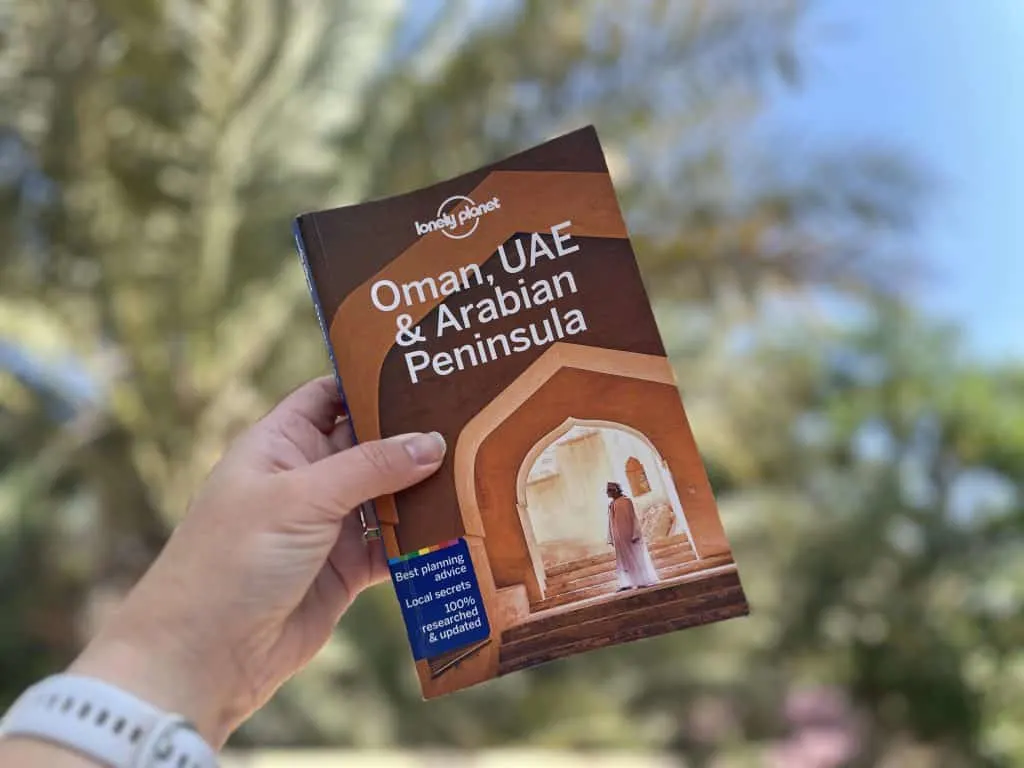 Copy of the Lonely Planet's Oman, UAE & Arabian Peninsular travel guide held by hand in front of palm tree and blue sky background