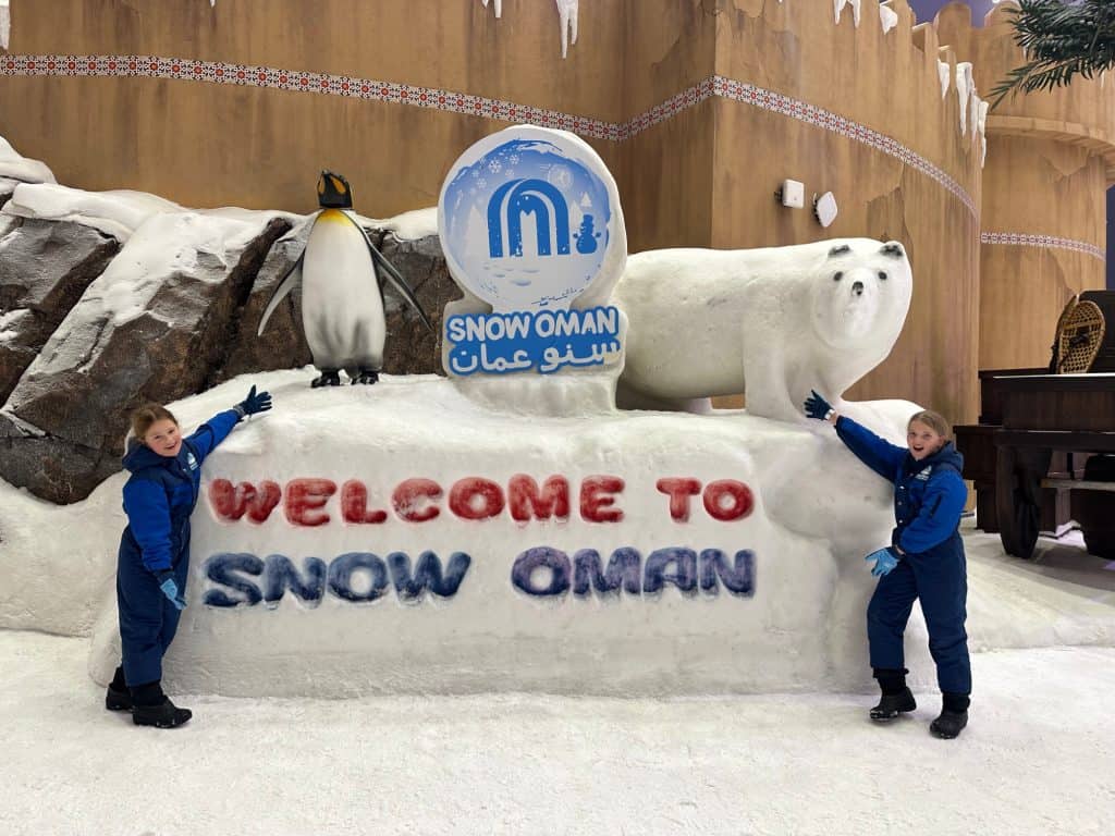 Children stand at either end of a 'Welcome to Snow Oman' sign on which a model penguin and polar bear stand