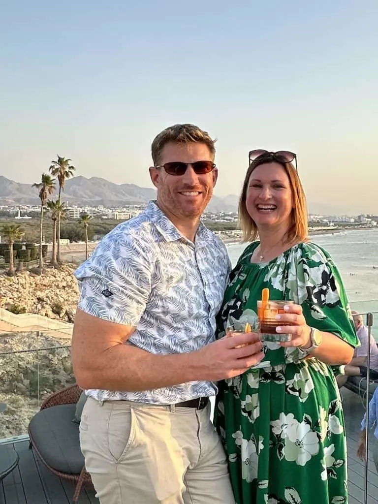 Claire and husband holding drink in front of a view of Qurum in Muscat at sunset. They are at Crown Plaza Muscat hotel