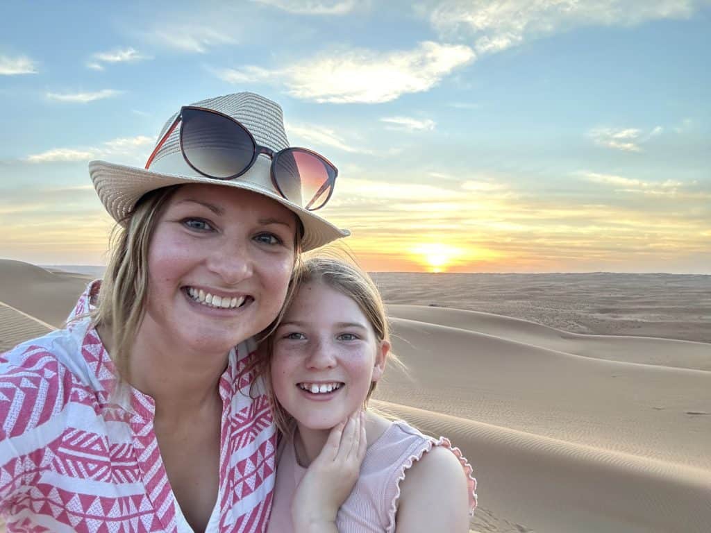 Claire and daughter stood on sand dune in Wahiba Sands desert while the sun sets behind them