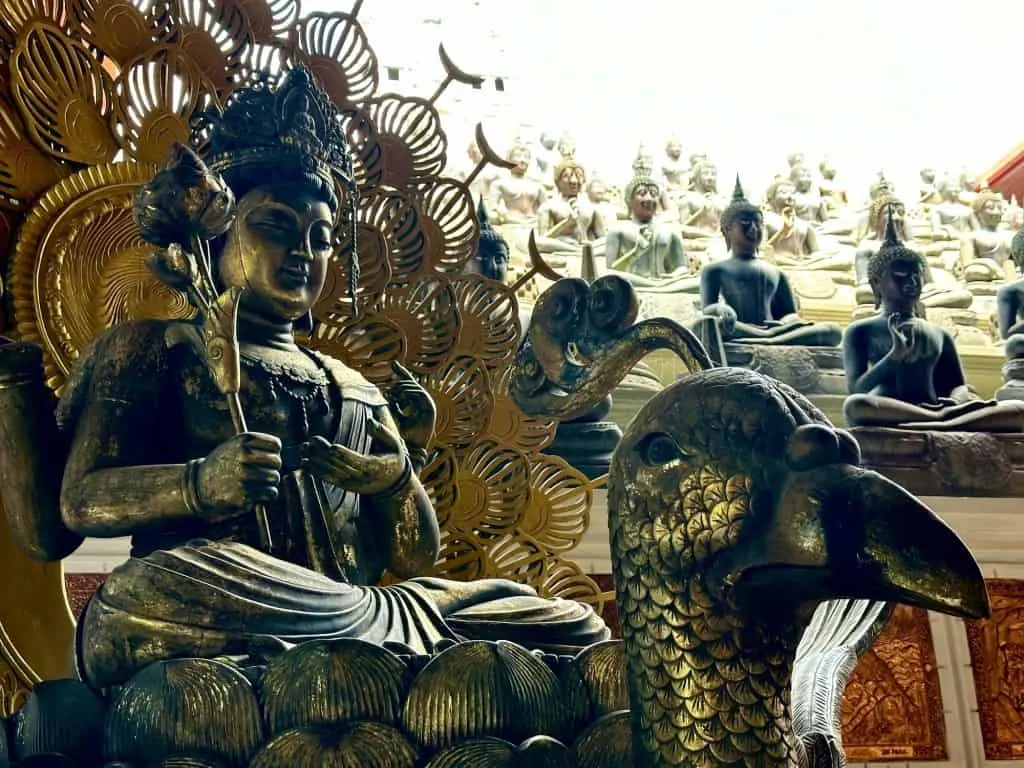 Brass Buddha on a peacock in front of dozens of Buddhas lined up inside temple