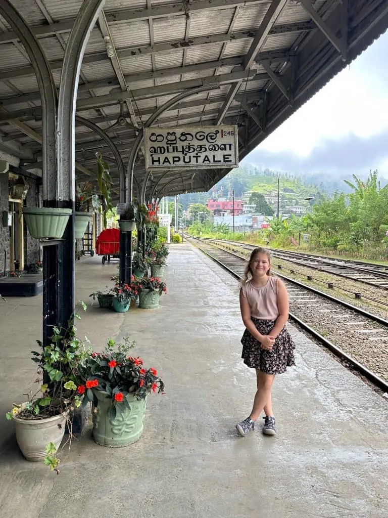 Our eight year old stood on the colonial built train station at Hapetule. The train tracks behind her are empty and there is a view of mountains behind her