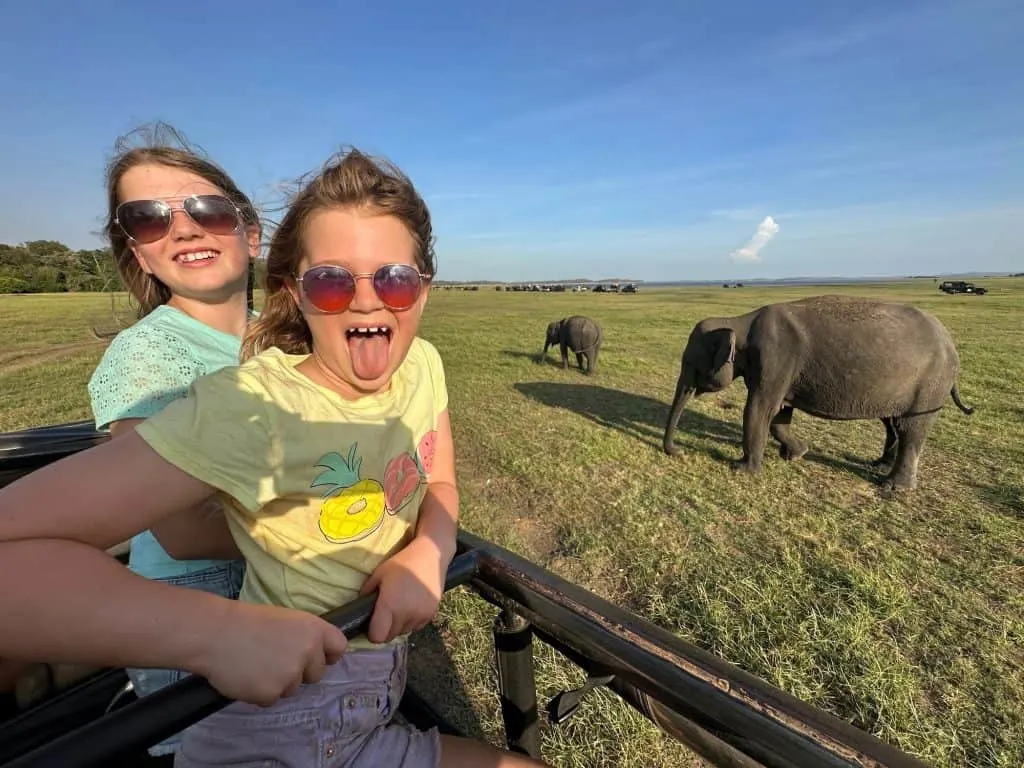Smiling girls pose in jeep with a family of elephants behind them