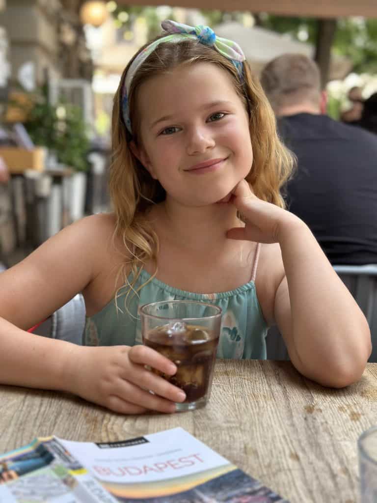 Our eight-year-old smiling at the camera while sat at a table in 0,75 Bistro in Budapest. She is holding a small glass of cola