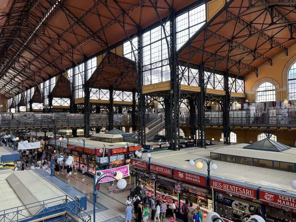 A view of the inside of Budapest's Central Market Hall which is an iron construction with large windows. There are stalls on the ground floor and a balcony of more stalls above.