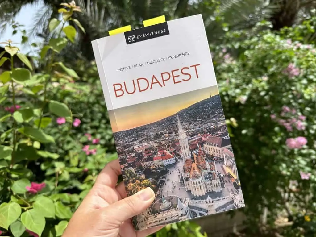 Claire's hand holds a copy of the Eye Witness Budapest guidebook in front of garden bushes and flowers