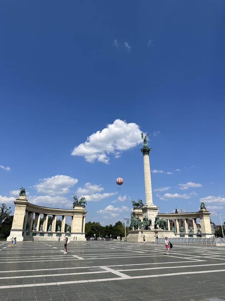 A column and statues in a large open square. In the background the sky is blue and a tethered hot air balloon can be seen in the sky