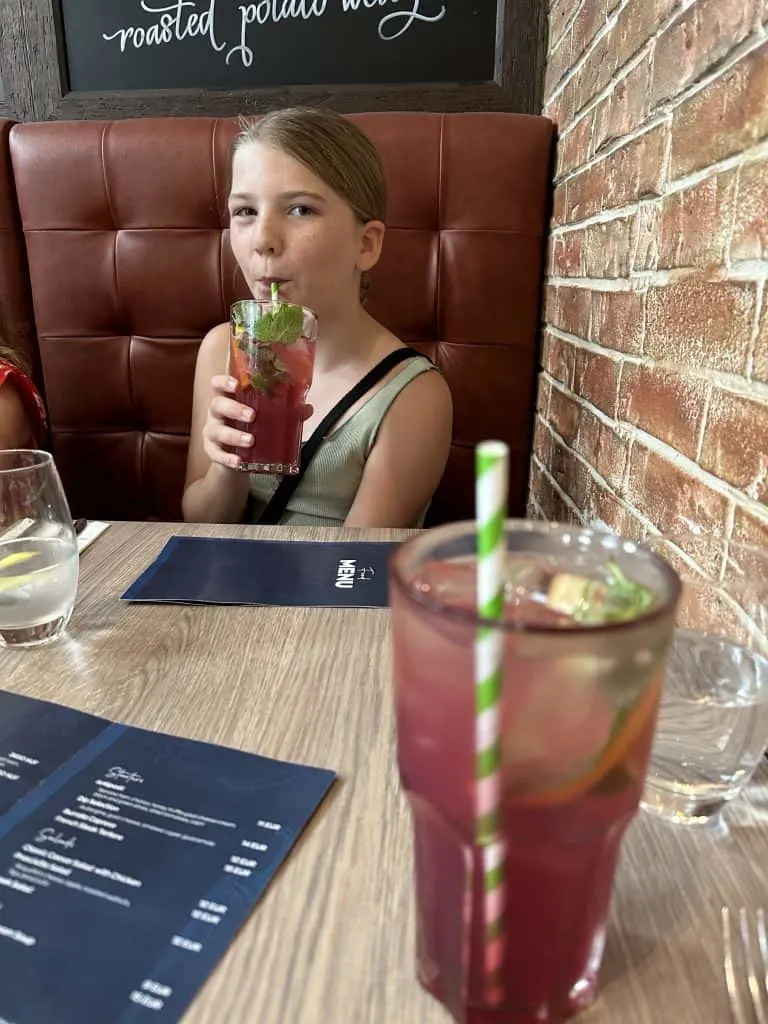 A raspberry lemonade on a restaurant table alongside a menu. Our 10-year-old daughter is drinking the same drink in the background